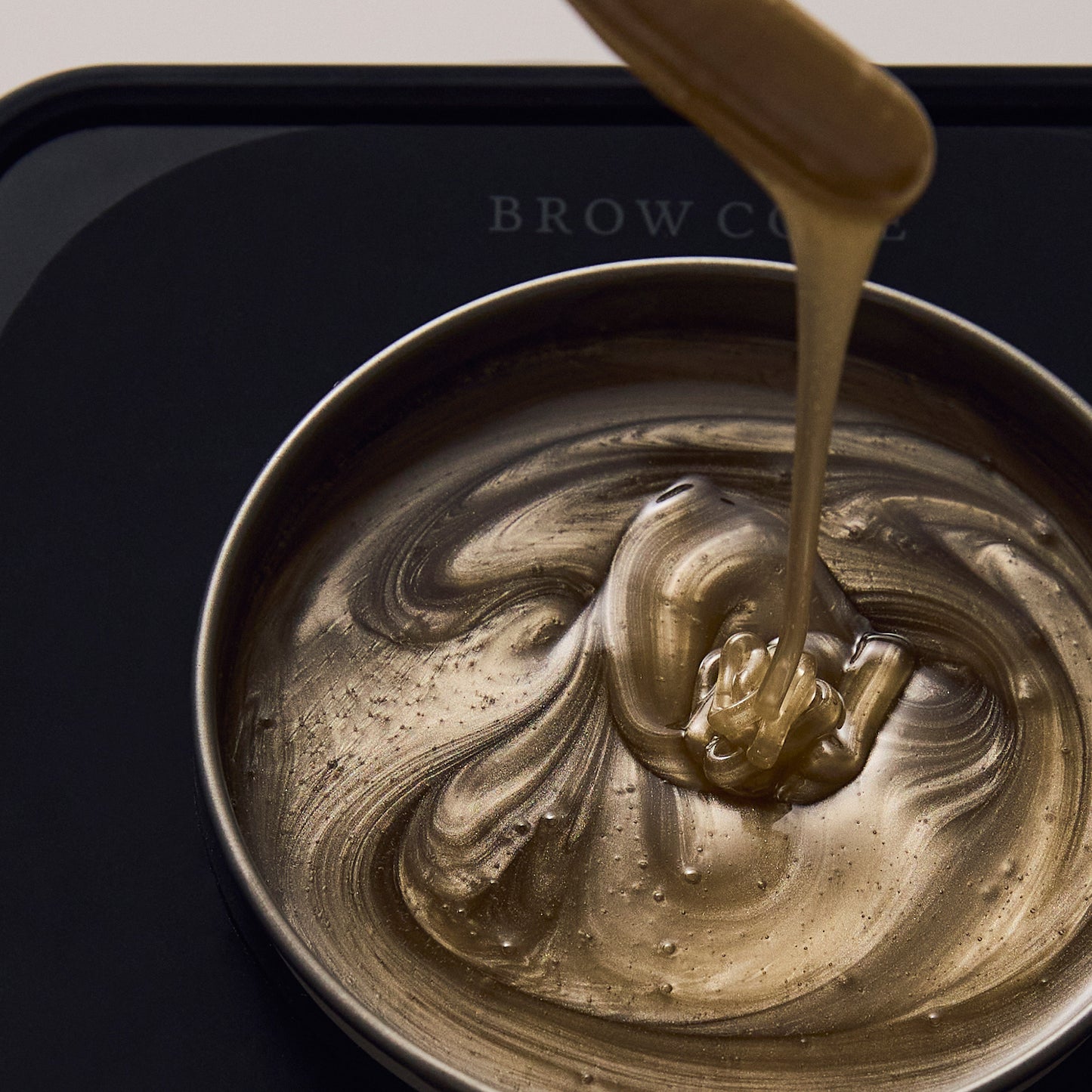 Close-up of Brow Code's Butter Bond wax being drizzled into a heated wax pot. The golden-brown wax has a smooth, creamy consistency, with the Brow Code branding subtly visible in the background