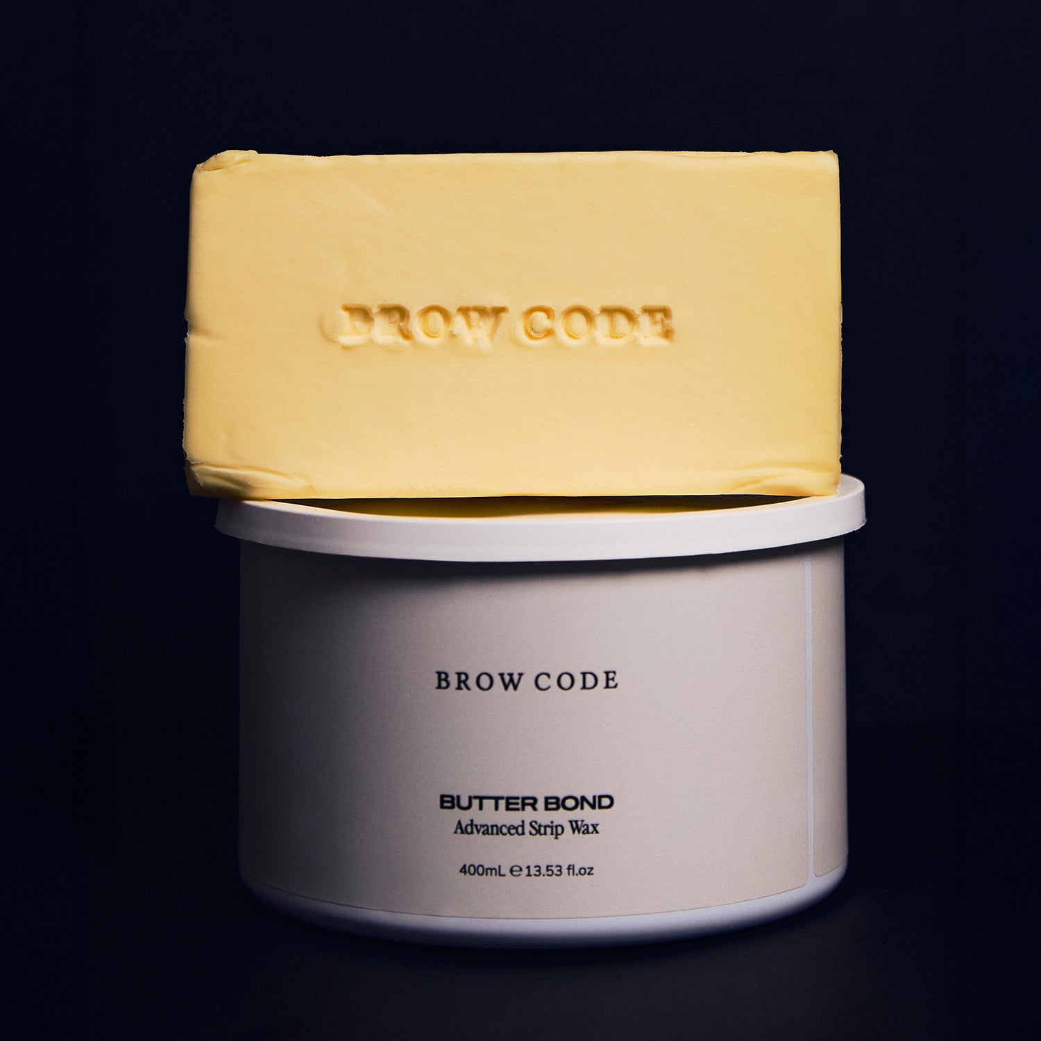 Close-up of a smooth, rectangular block of Butter Bond wax in its solid form, set against a dark navy background. The creamy yellow tone and clean edges highlight its buttery, hypoallergenic formula