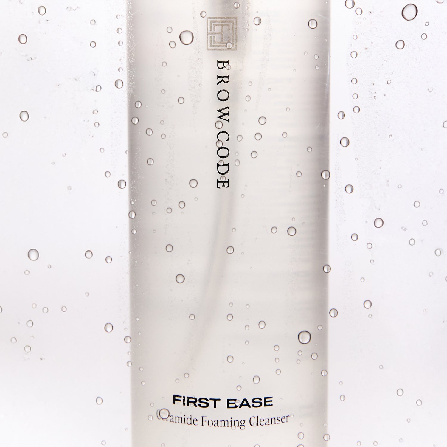 Brow Code First Base Ceramide Foaming Cleanser bottle with vertical logo, shown close-up behind a misted surface with water droplets, on a white background