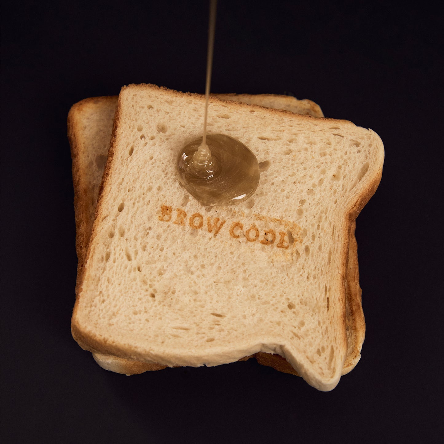 Butter Bond wax dripping onto a piece of toast embossed with the Brow Code logo. The rich, golden wax mimics melted butter, playfully highlighting the wax's buttery texture against the crisp, stamped surface of the toast.