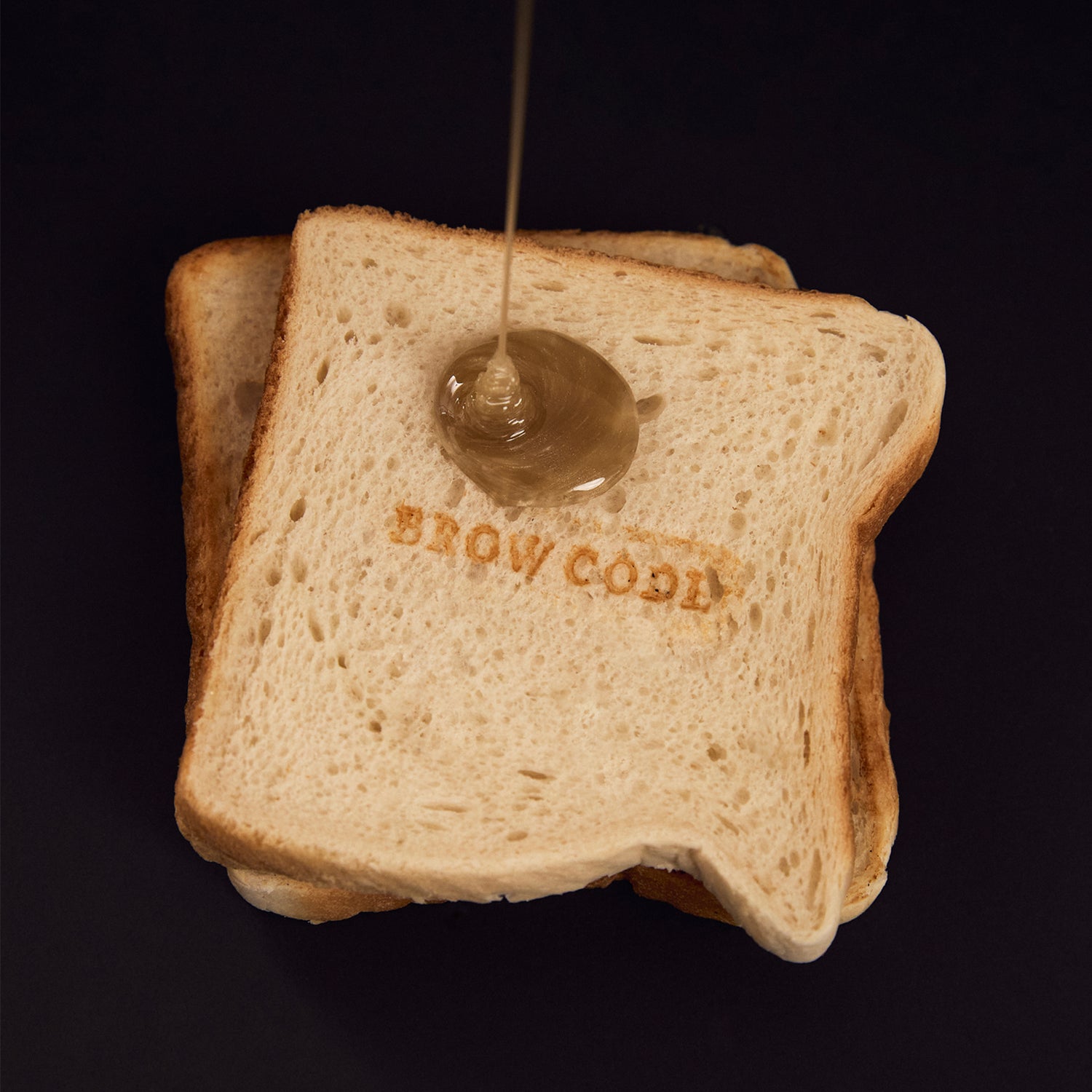Butter Bond wax dripping onto a piece of toast embossed with the Brow Code logo. The rich, golden wax mimics melted butter, playfully highlighting the wax's buttery texture against the crisp, stamped surface of the toast.