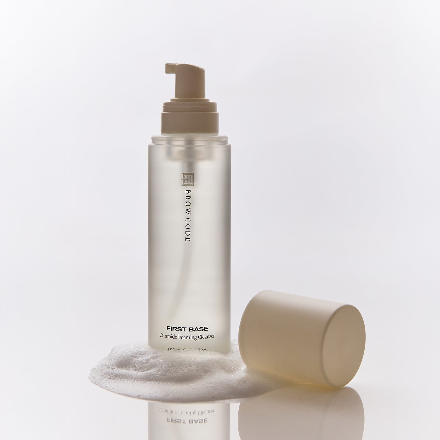 Brow Code First Base Ceramide Foaming Cleanser bottle with beige pump, cap beside it and foam around the base on a white background