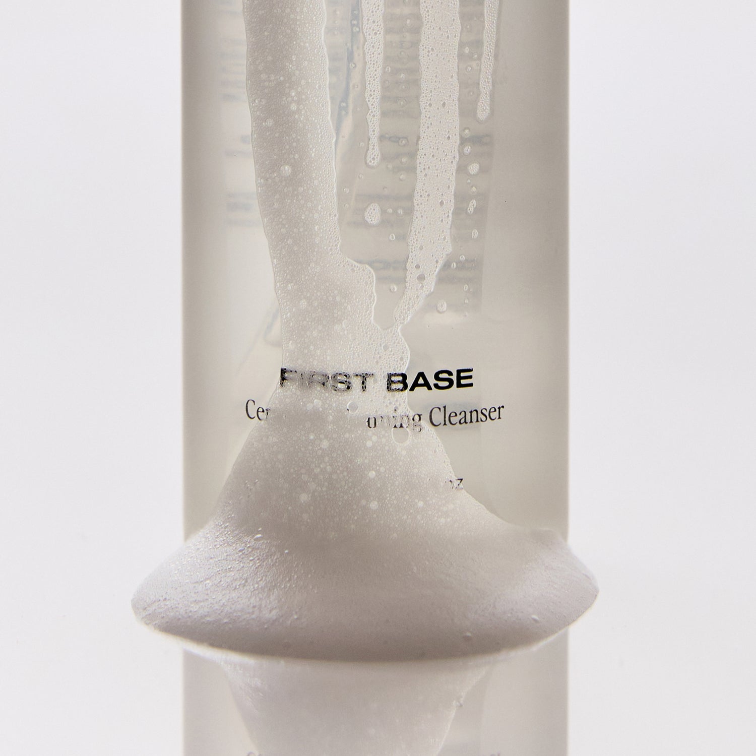 Brow Code First Base Ceramide Foaming Cleanser close-up of bottle with foam cascading down the front, partially covering the label, on a white background