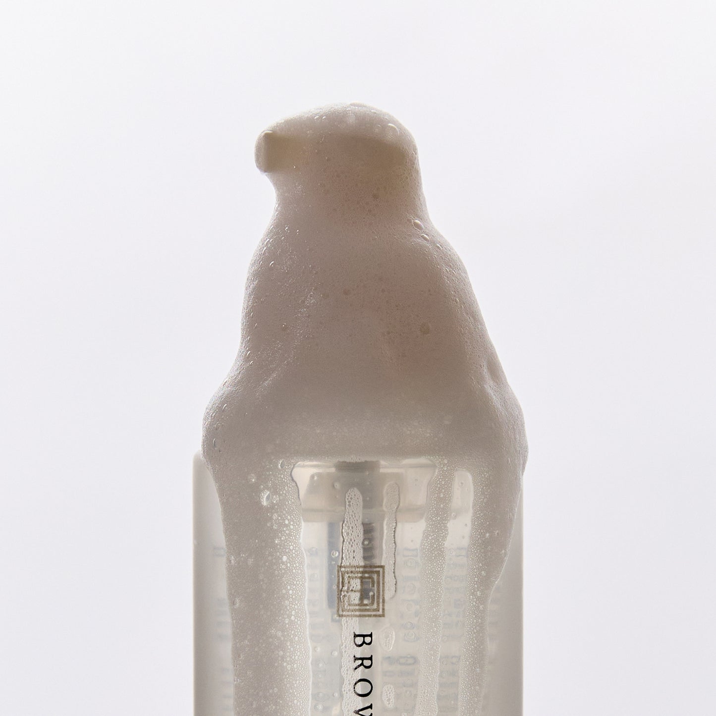 Brow Code First Base Ceramide Foaming Cleanser bottle with beige pump, covered in thick foam forming a dome over the top, on a white background